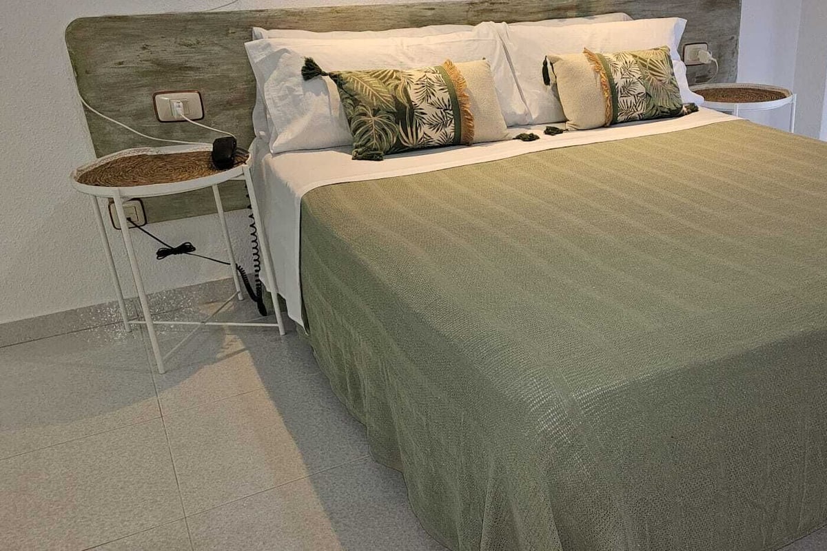 Una cama bien hecha con una colcha verde y varias almohadas, una mesita de noche con una lámpara y un enchufe en la pared.