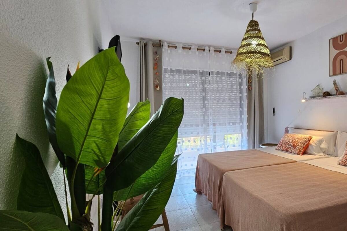 A hotel room with two beds, a large plant, a hanging light fixture, and a view of the outdoors.