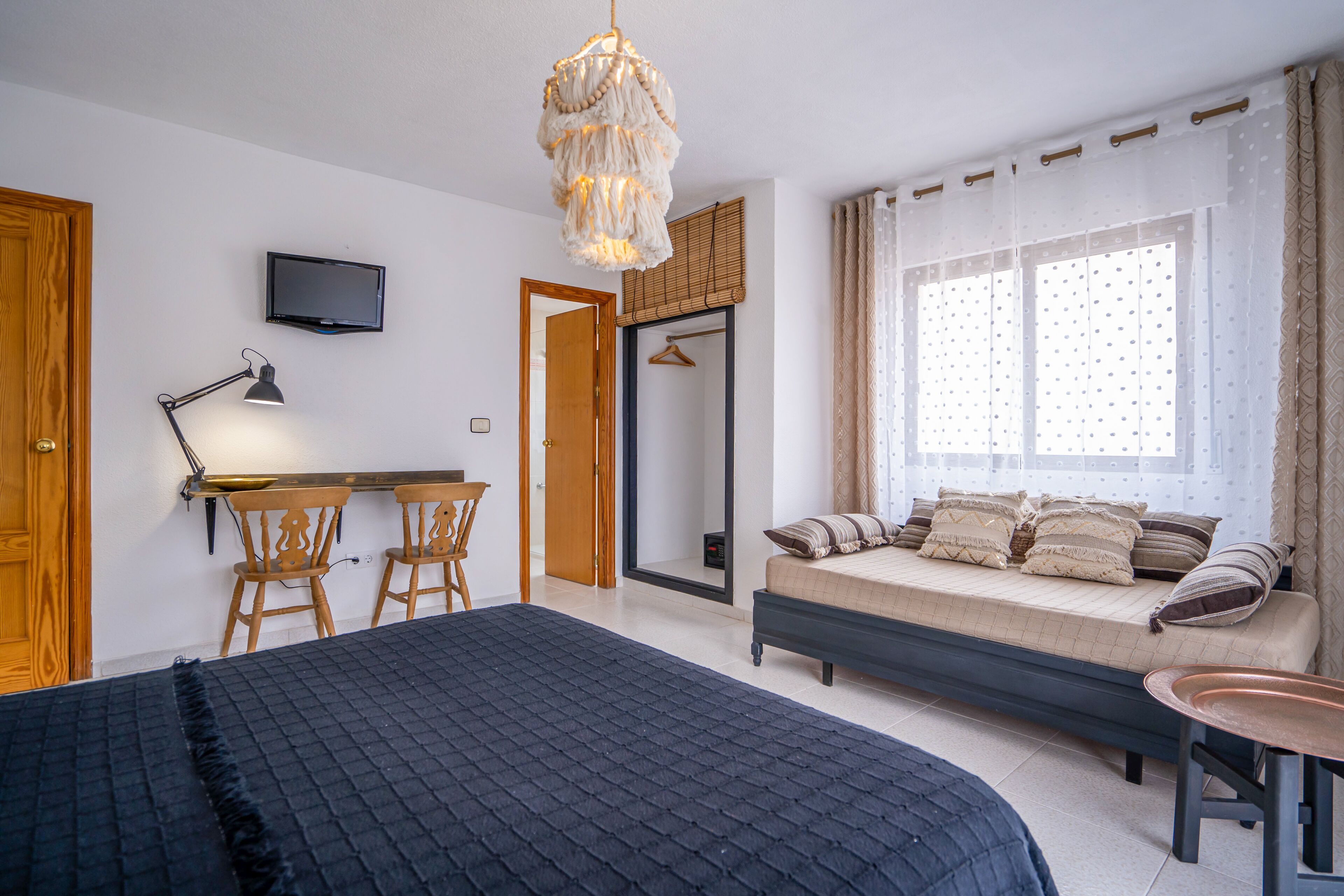 A bedroom with a bed, a desk, two chairs, a TV, and a window with curtains.