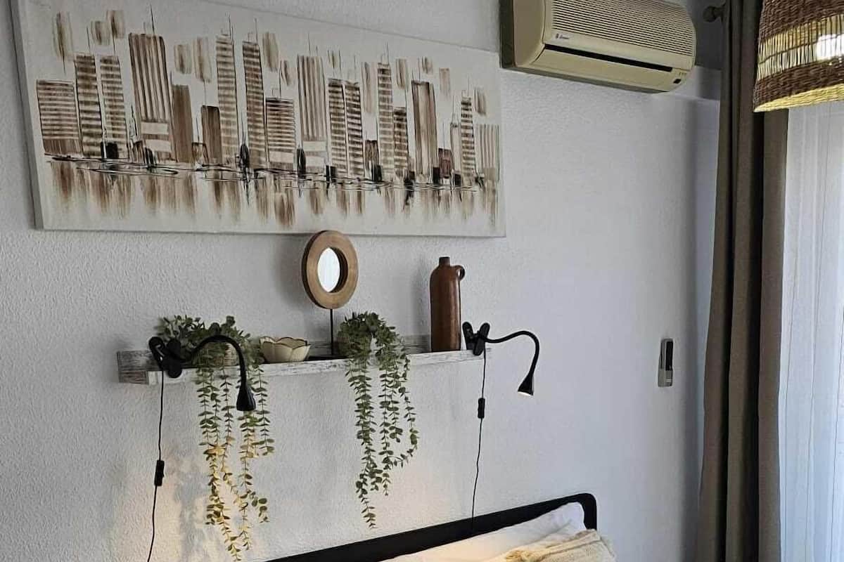 Una cama bien hecha con un edredón estampado, un aire acondicionado de pared, un adorno en la pared y una planta colgante.
