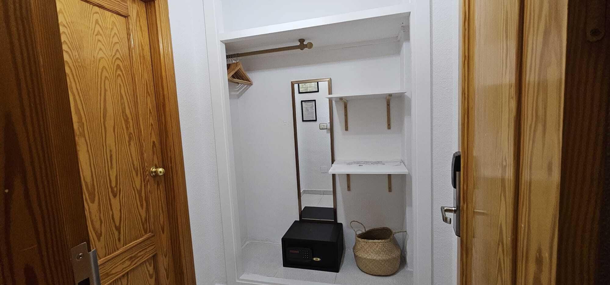A small, narrow hallway with a wooden door on the left, a white wall with a shelf and a small black box, and a cat tree on the right.