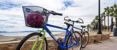 A blue bicycle with a basket and a pink helmet on the front wheel, parked on a brick path near a beach with palm trees and a stone wall.