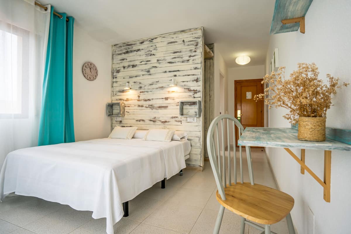 Un dormitorio con cama, una silla, una mesita con un florero y un reloj de pared.
