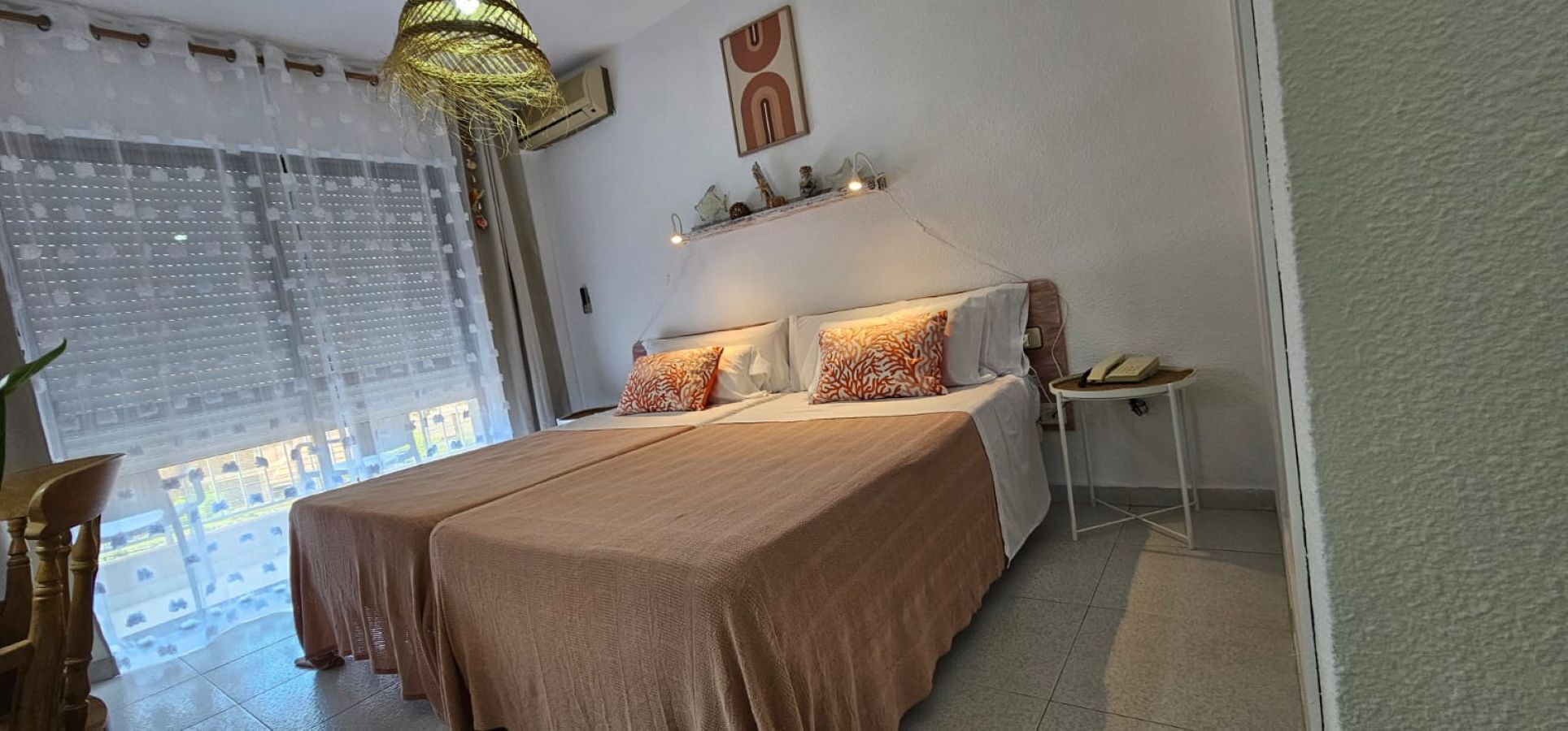 A bedroom with a large bed, two pillows, a bedside table with a lamp, and a window with sheer curtains.