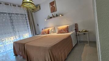A bedroom with a large bed, two pillows, a bedside table with a lamp, and a window with sheer curtains.