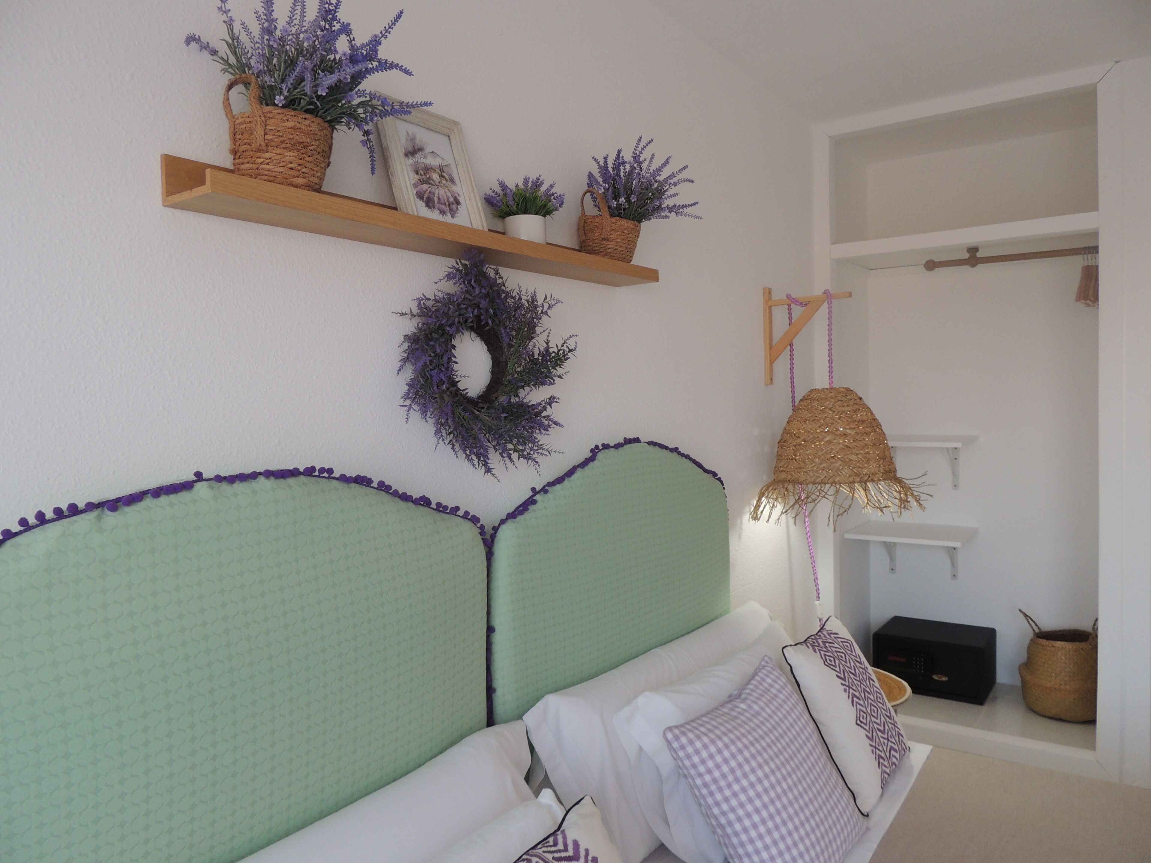 A room with a green upholstered headboard, a hanging lamp, a shelf with plants, and a wreath on the wall.