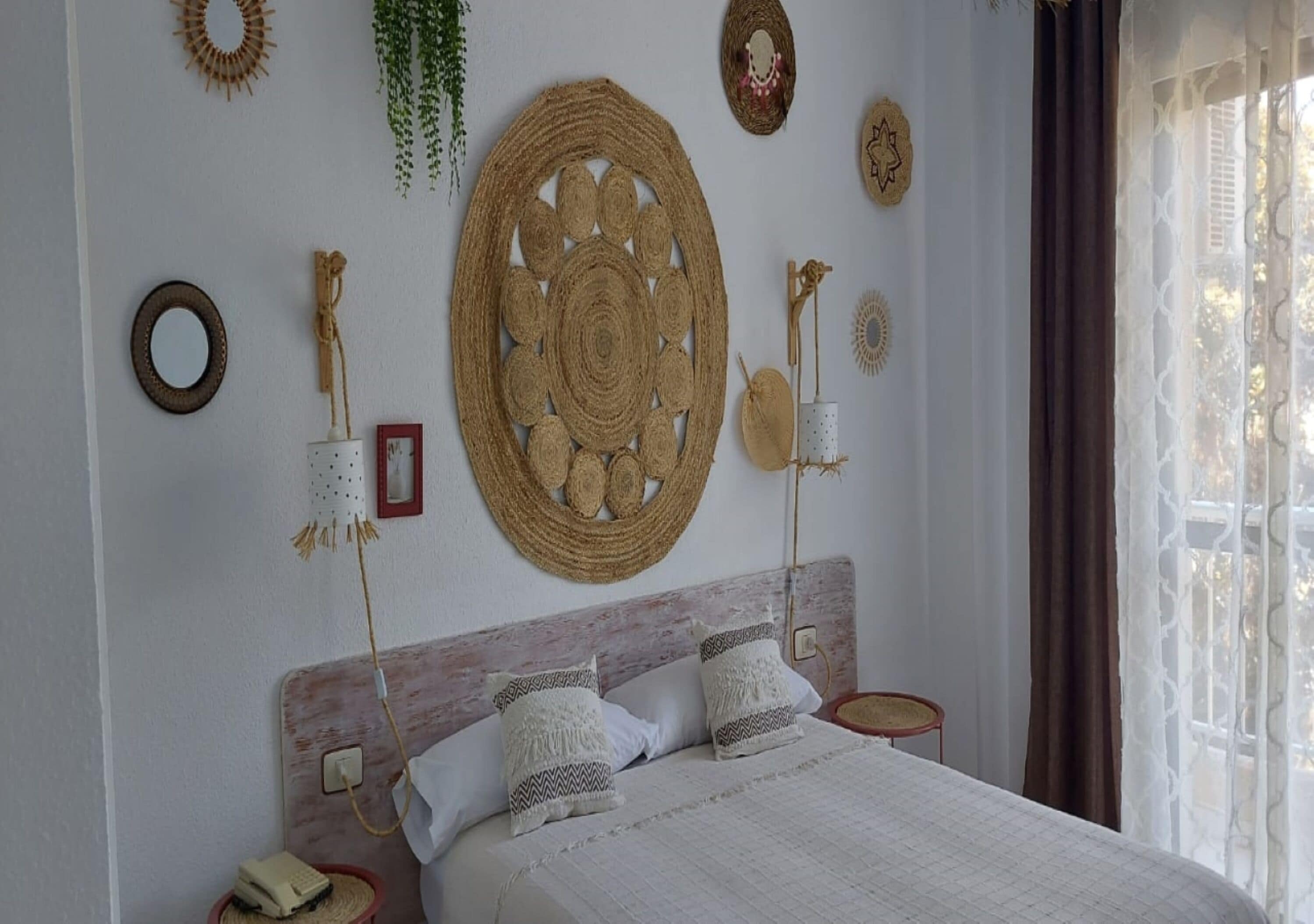Une chambre à coucher comprenant un lit, deux tables de chevet, un téléphone fixé au mur et divers objets décoratifs, notamment des éléments de décoration murale tissés et une plante suspendue.