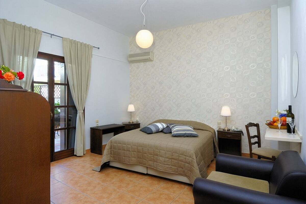 A bedroom with a bed, a desk, a chair, a small table with a lamp, and a window with curtains.