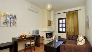 A living room with a brown sofa, a coffee table, a TV on a wooden stand, a fireplace, and artwork on the wall. - Enalion Hotel (South Pelion)