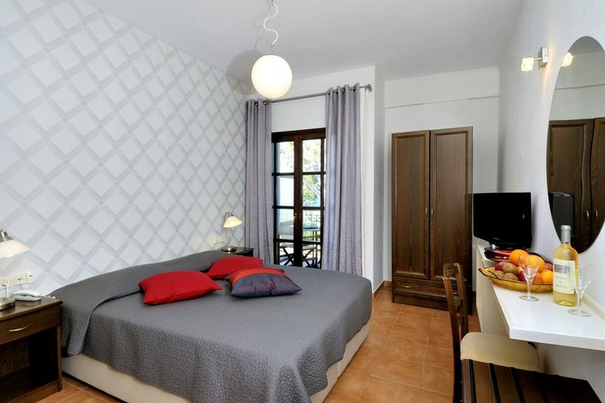 A bedroom with a bed, bedside table, lamp, curtains, window, wardrobe, TV, and a breakfast area with fruit and wine.