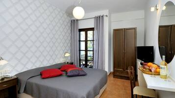 Quarto com cama, mesa de cabeceira, luminária, cortinas, janela, guarda-roupa, televisão e um espaço de refeições com frutas e vinho.