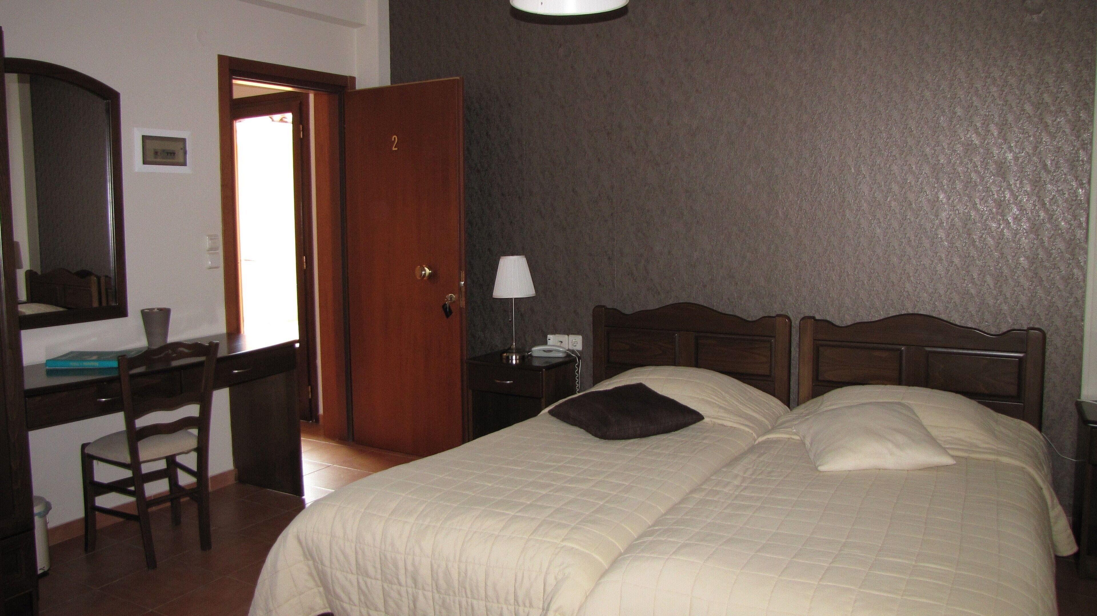 Habitación de hotel con cama, escritorio, silla, mesita de noche con lámpara y puerta.