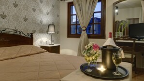 A hotel room with a bed, a bedside table with a lamp, a window with curtains, a tray with a wine glass, and a television. - Enalion Hotel (South Pelion)