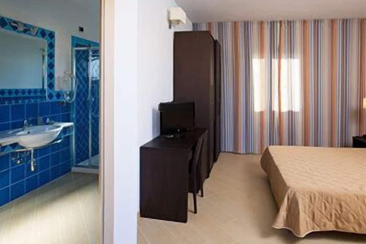 Quarto de hotel com banheiro, cama, escrivaninha e televisão.