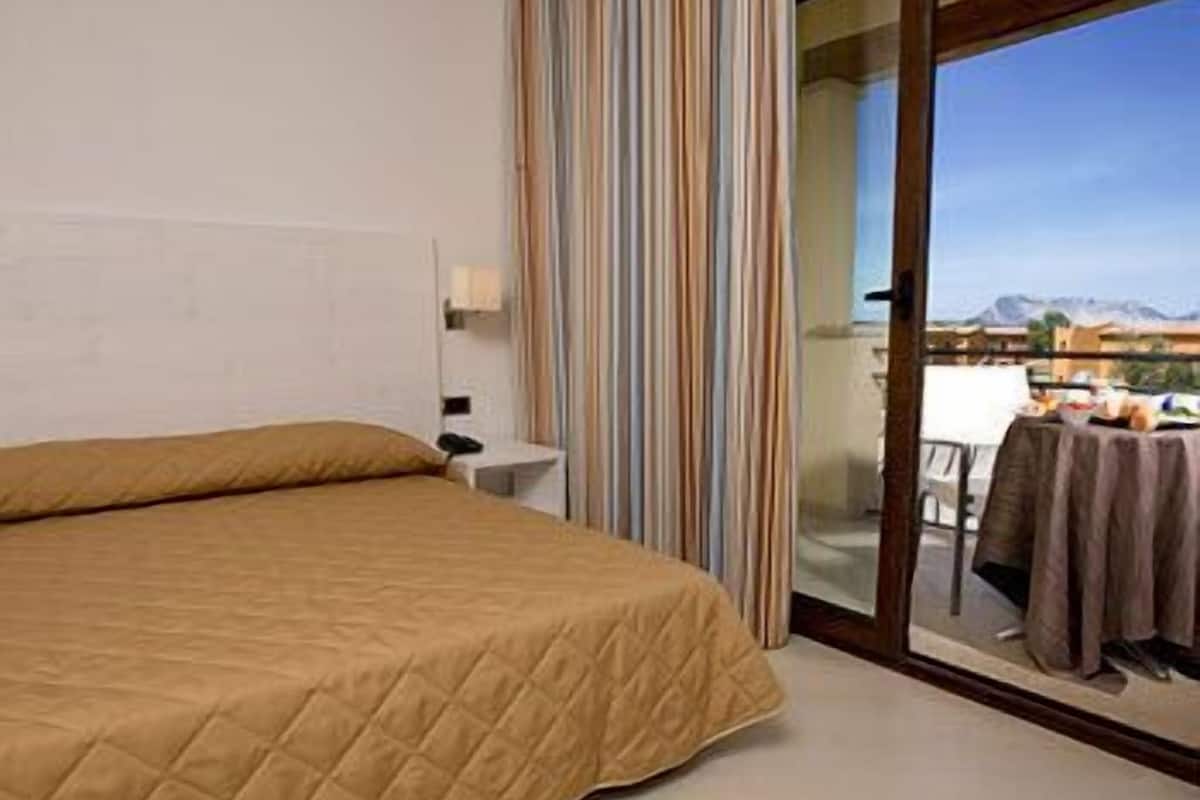 Quarto de hotel com cama, mesa de cabeceira, janela com cortinas e vista para uma varanda com mesa e cadeiras.