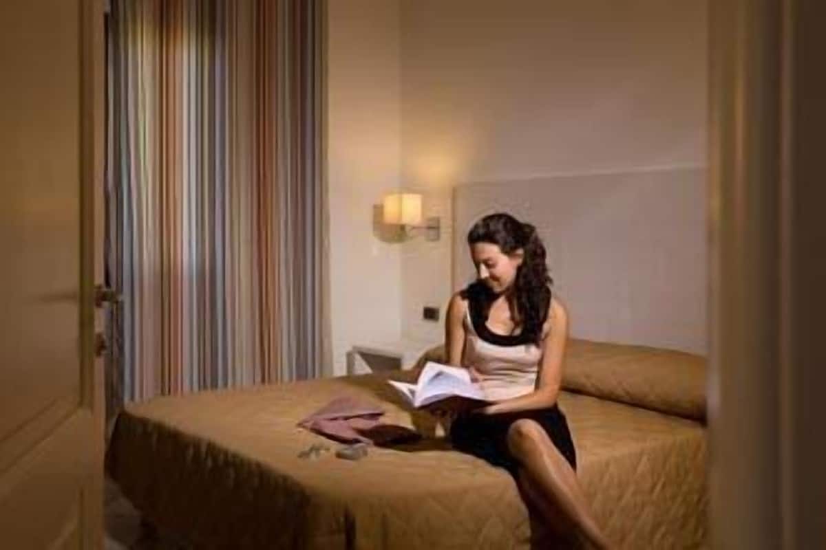 Uma pessoa sentada na cama em um quarto com cortina listrada e mesa de cabeceira com abajur.