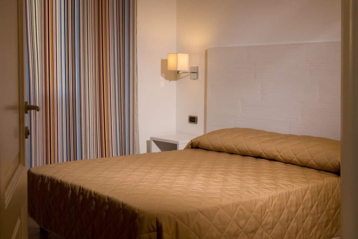 Quarto de hotel com cama, mesa de cabeceira, luminária na parede e cortina listrada.