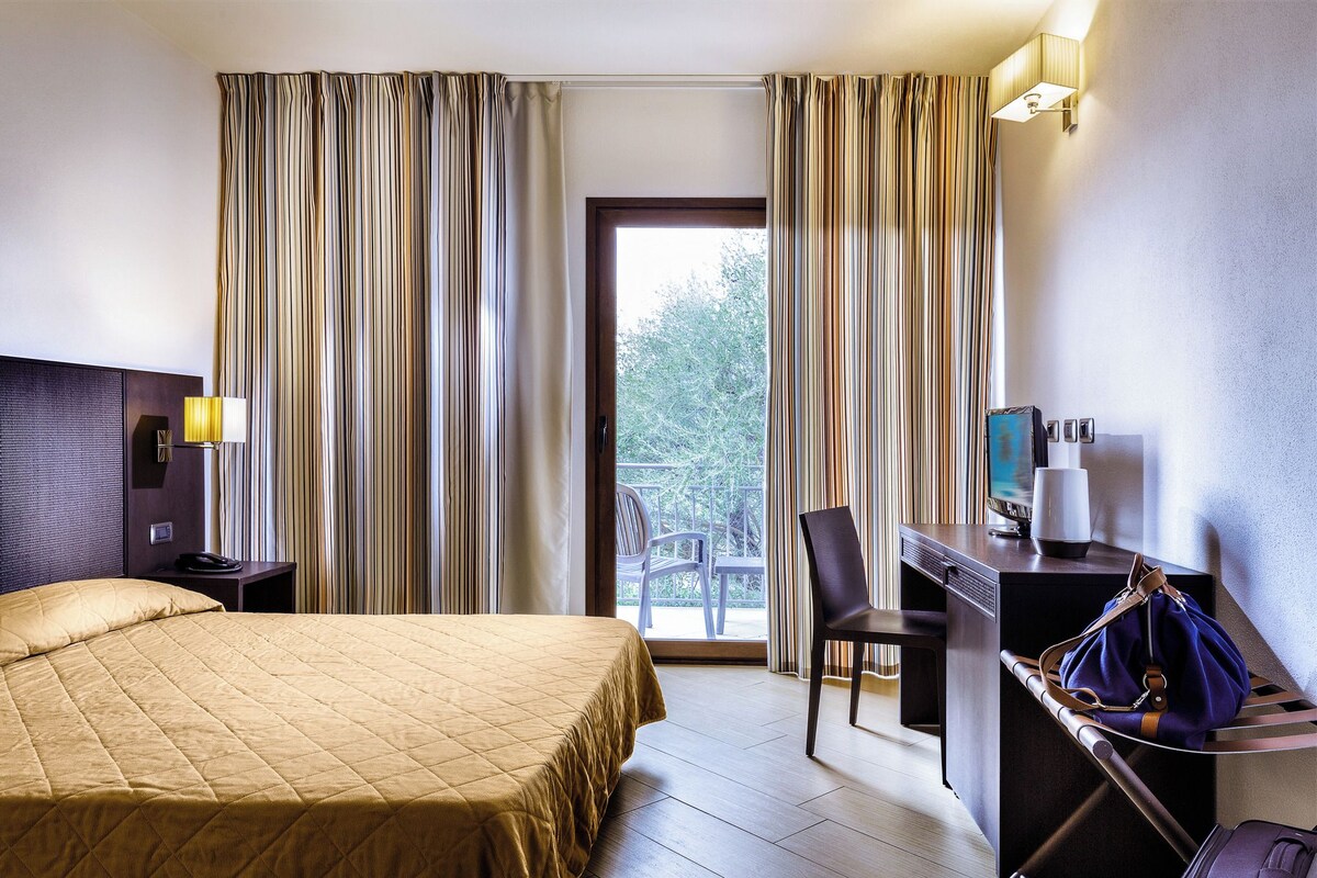 Habitación de hotel con cama, escritorio, silla, lámpara y vista al exterior.
