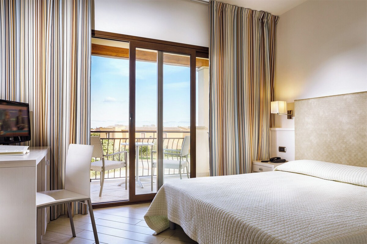 Habitación de hotel con cama, escritorio con televisor, silla, balcón con vistas y cortinas.