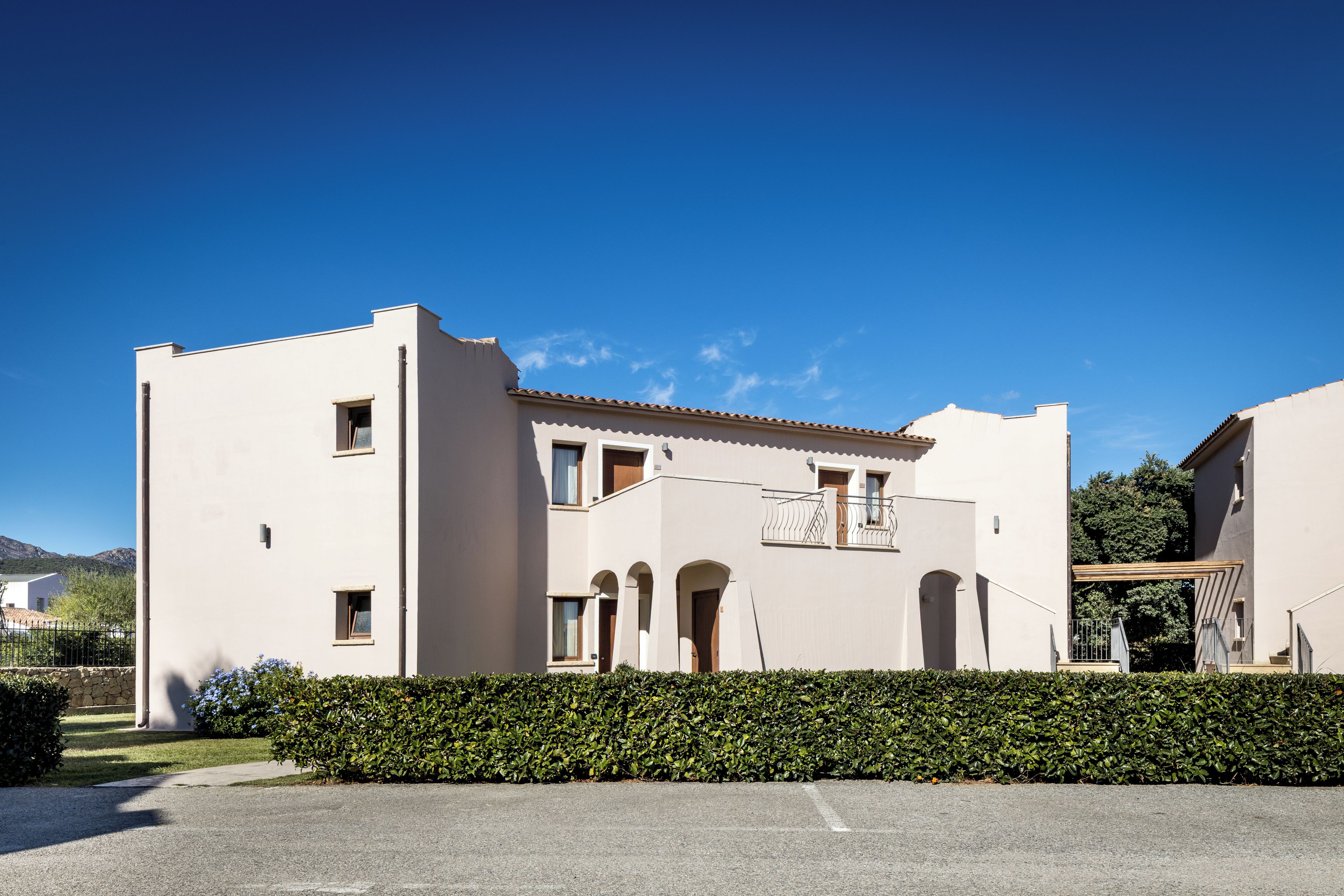 A two-story building with a flat roof, a balcony, and a hedge in front.