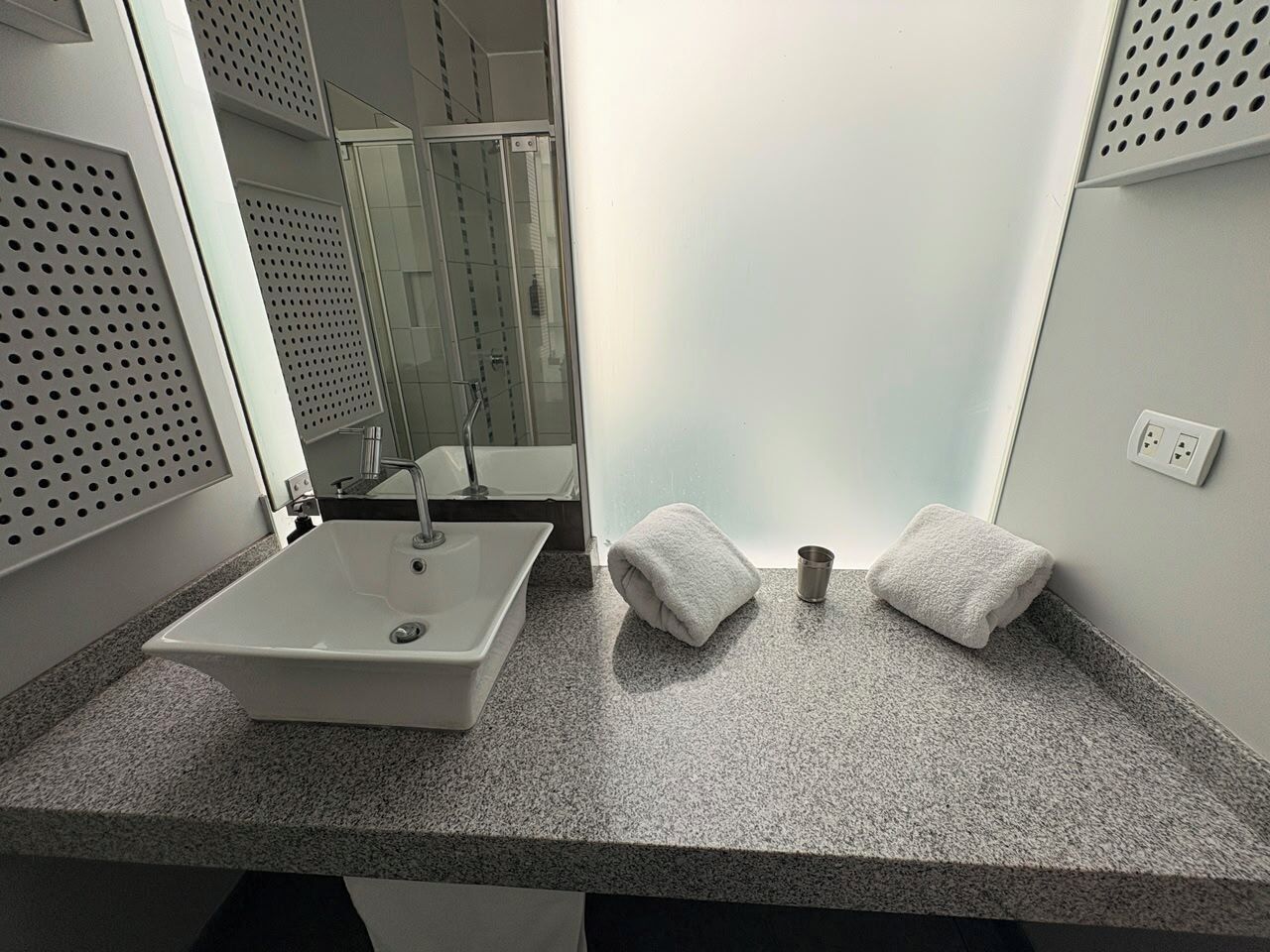 A bathroom with a white sink, two towels, a cup, and a glass shower enclosure.