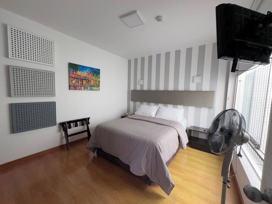 Un dormitorio con cama, un banco, un escritorio, un ventilador y un cuadro en la pared.