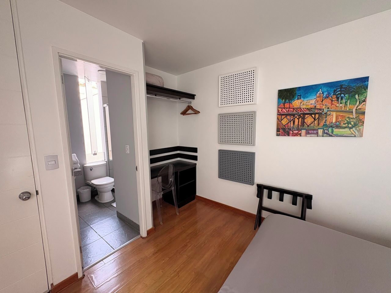 A room with a wooden floor, a bed, a chair, a desk, a hanging coat rack, a painting, and a bathroom with a toilet and sink.