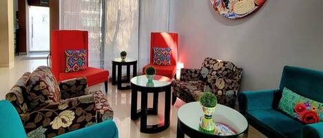 Una sala de estar moderna con sofás color turquesa, sillas rojas y mesas negras. Hay ventanas amplias con cortinas translúcidas, una lámpara colgante roja y obras de arte en las paredes.