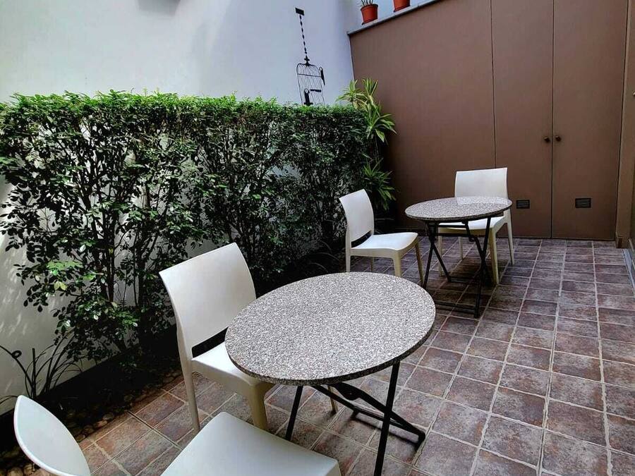 Un área de patio con dos mesas redondas y cuatro sillas, rodeada por un seto bajo y plantas en macetas.