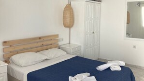 Un dormitorio con cabecera de madera, mesita de noche blanca, armario blanco, colcha azul y dos toallas enrolladas sobre la cama.