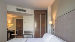A hotel room with a bed, a desk, a chair, a wardrobe, and a wall-mounted lamp. - Hotel Infanta Leonor (Ecija)