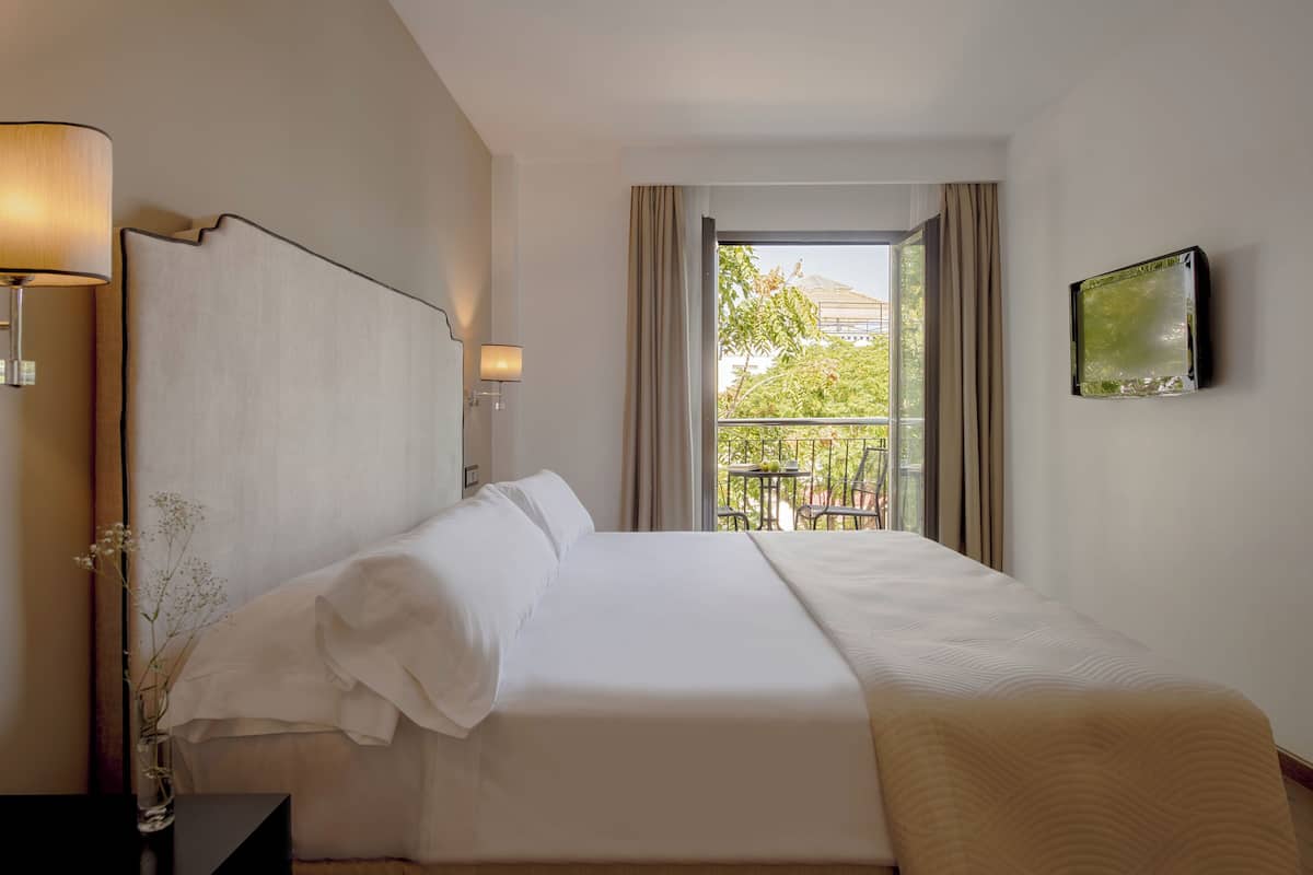 Una habitación de hotel con una cama grande, una mesita de noche con lámpara, una ventana con cortinas y un cuadro en la pared.
