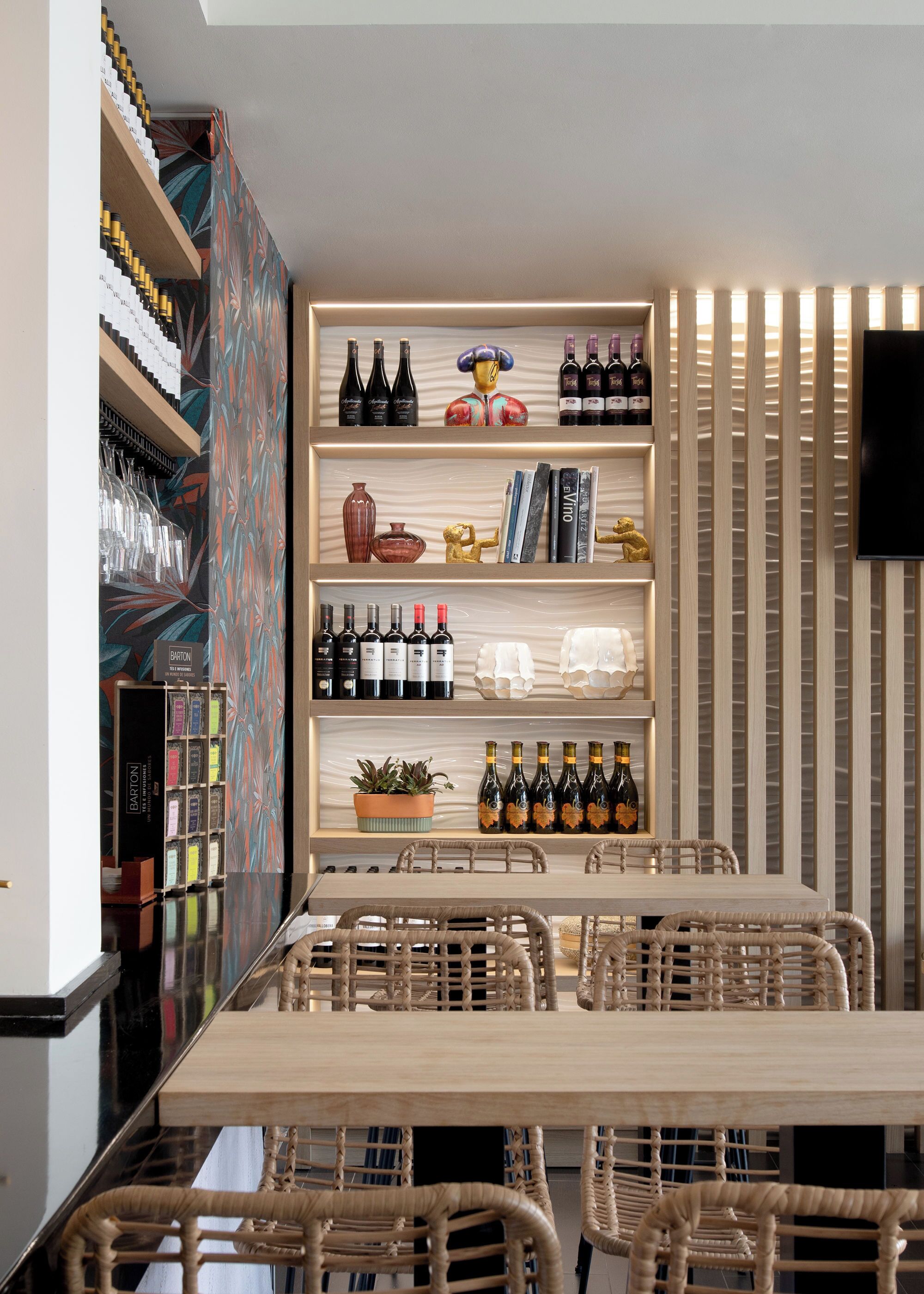 A modern interior with a long wooden table and wicker chairs, surrounded by shelves with wine bottles and decorative items.