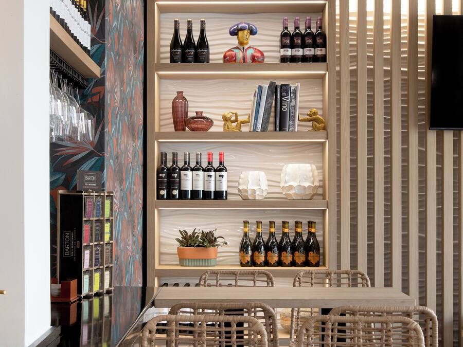 Un interior moderno con una larga mesa de madera y sillas de mimbre, rodeado de estanterías con botellas de vino y objetos decorativos.