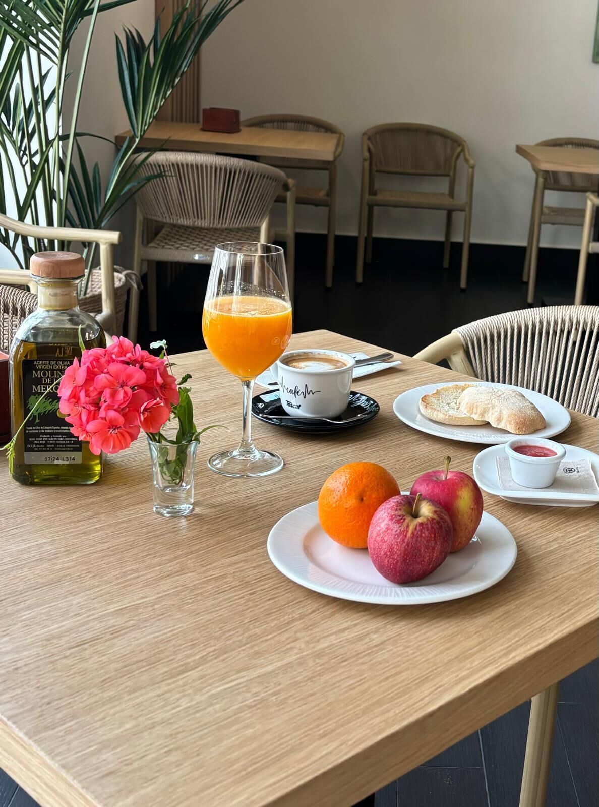 Una mesa de desayuno con una botella de aceite de oliva, un vaso de zumo de naranja, una taza de café, un plato con dos manzanas y un plato de pan con mermelada.