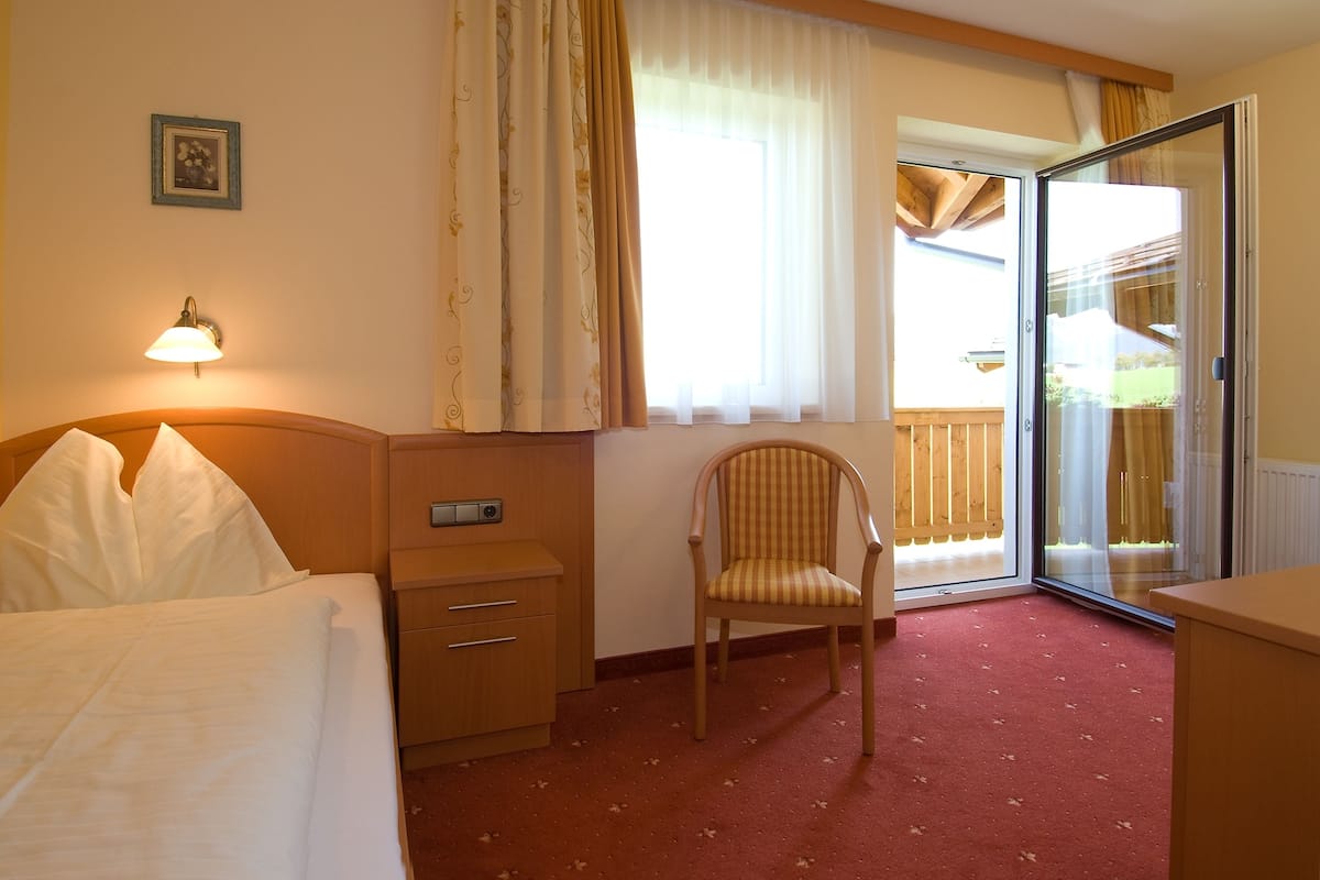 Une chambre d’hôtel comprenant un lit, une chaise, un bureau avec une lampe, une fenêtre avec des rideaux et une porte-fenêtre coulissante donnant sur un balcon.