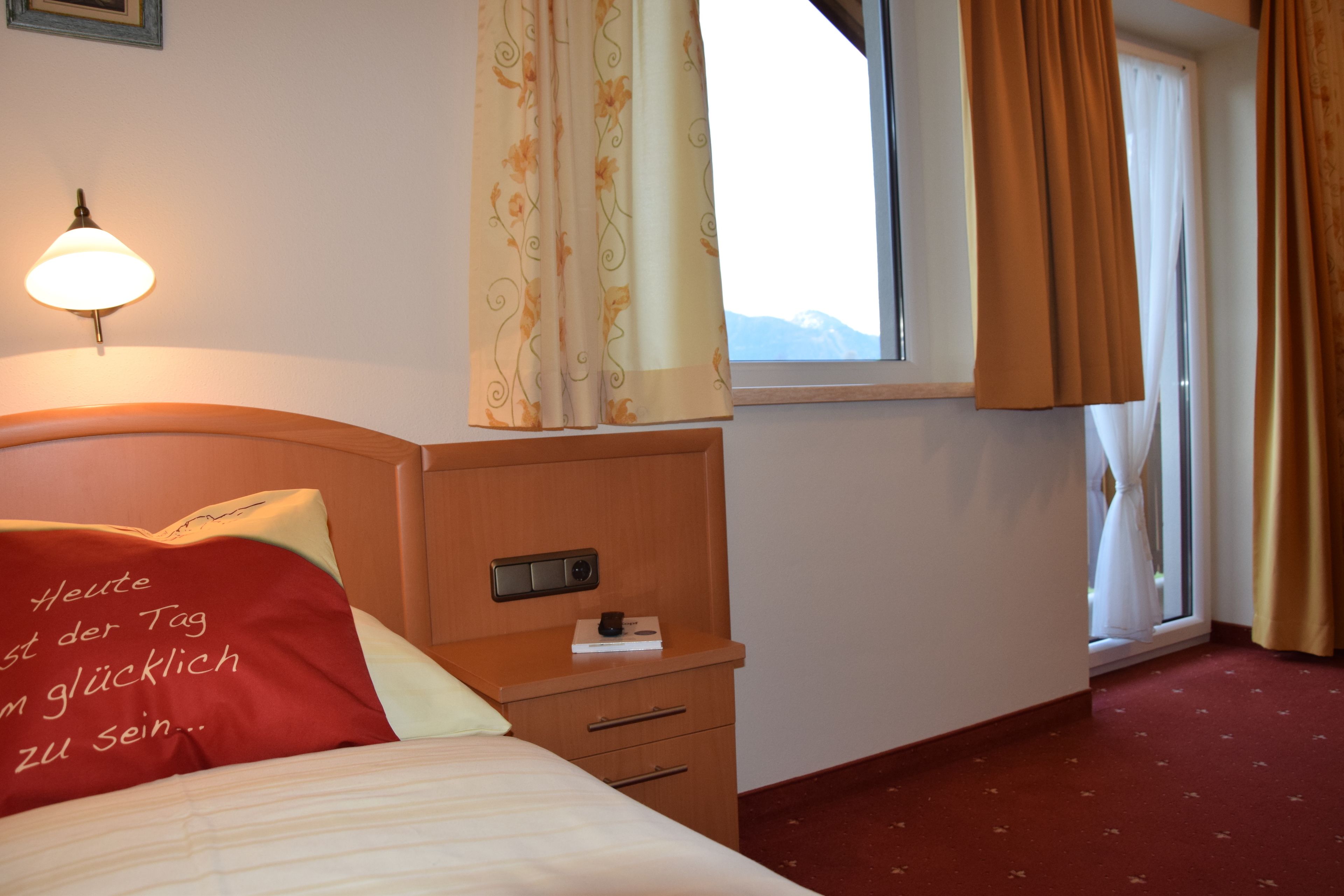 Habitación de hotel con cama, mesita de noche con lámpara, ventana con cortinas y vistas a las montañas.