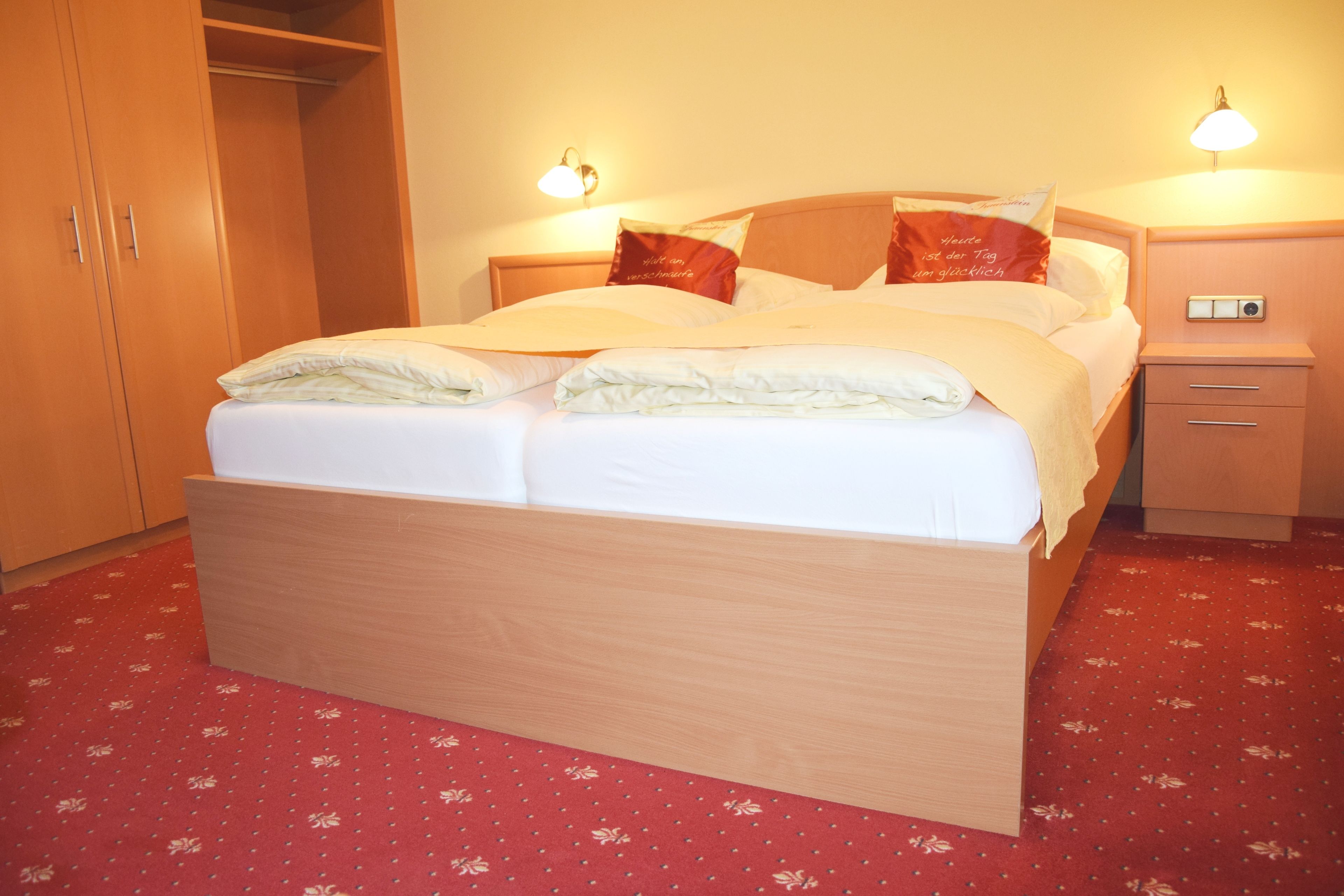 A neatly made bed with white linens and two pillows, a wooden headboard, a red carpet with a floral pattern, and a nightstand with a lamp.