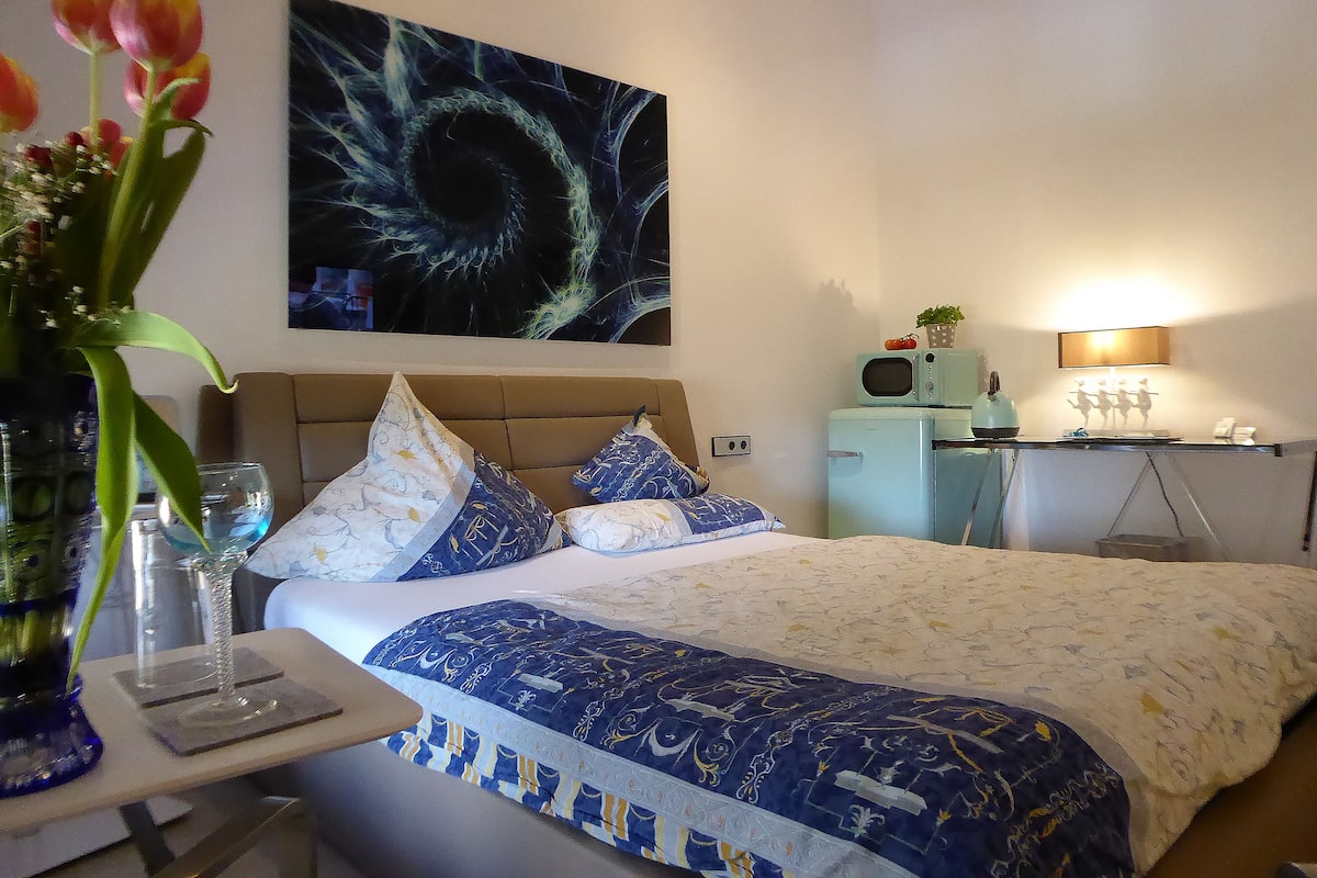 A bedroom with a bed, a desk, a lamp, and a painting on the wall.