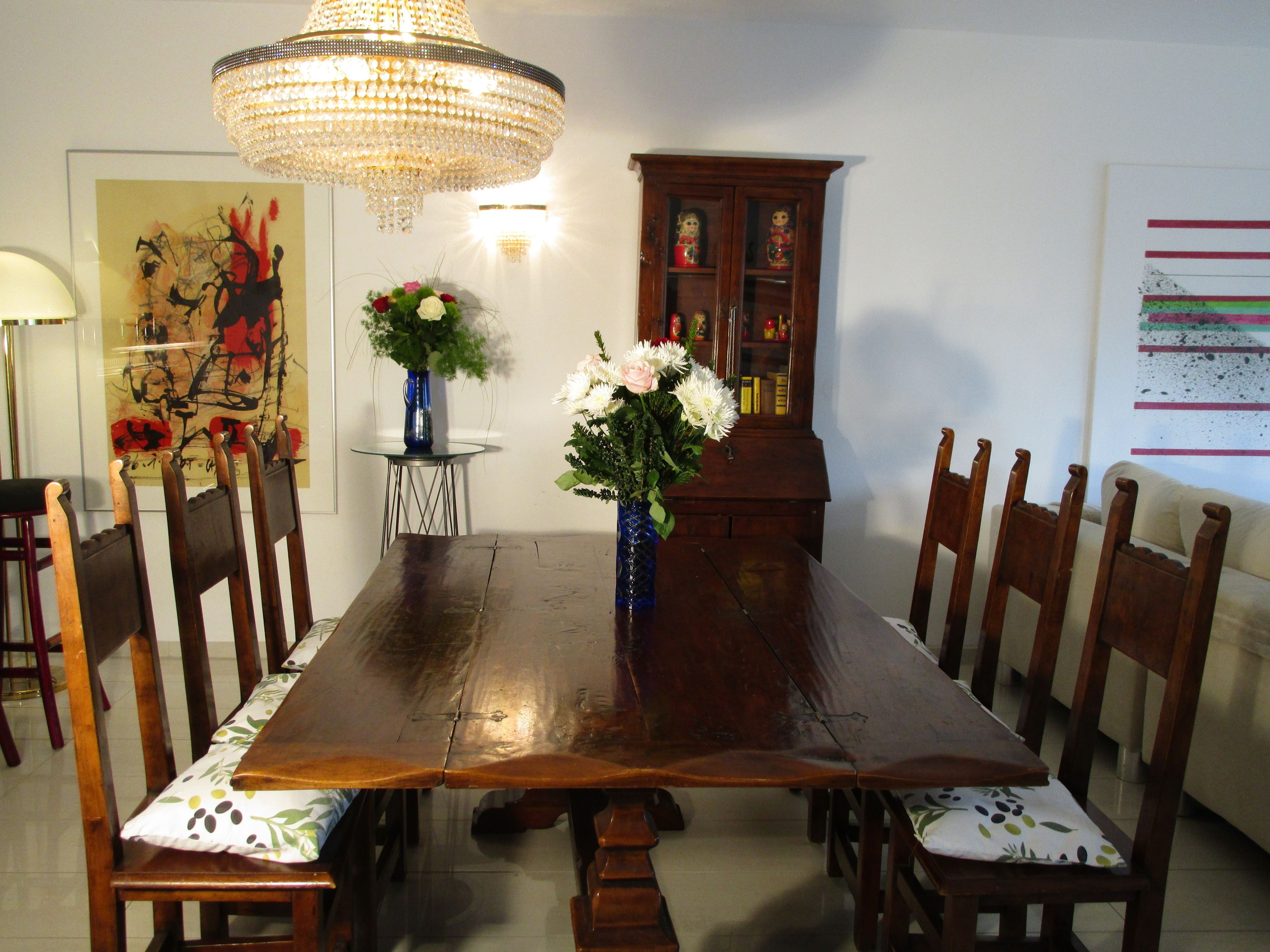 A dining room with a wooden table, chairs, a chandelier, a vase with flowers, and a cabinet.
