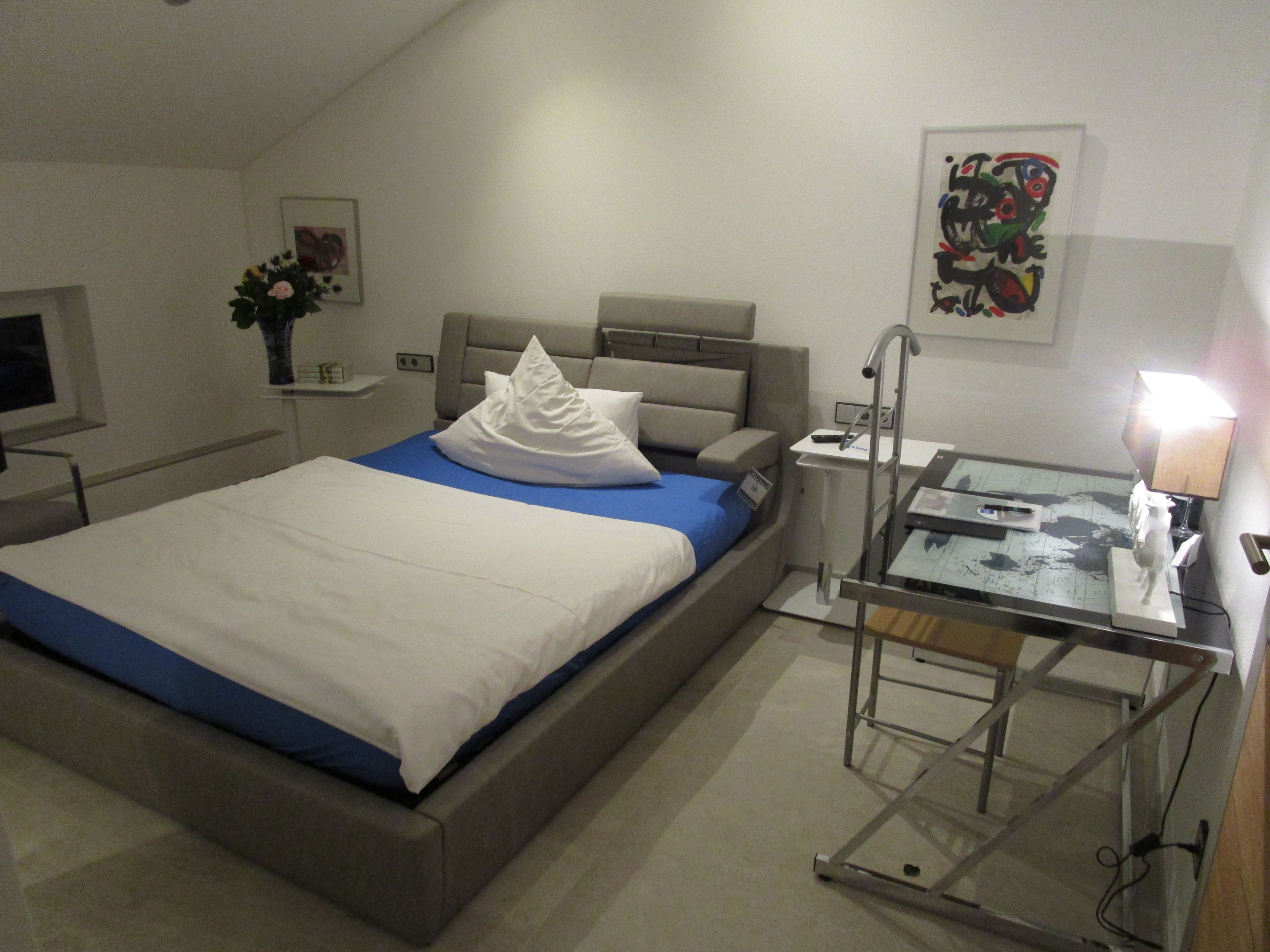 A hotel room with a bed, bedside table, lamp, and a small desk with a chair.