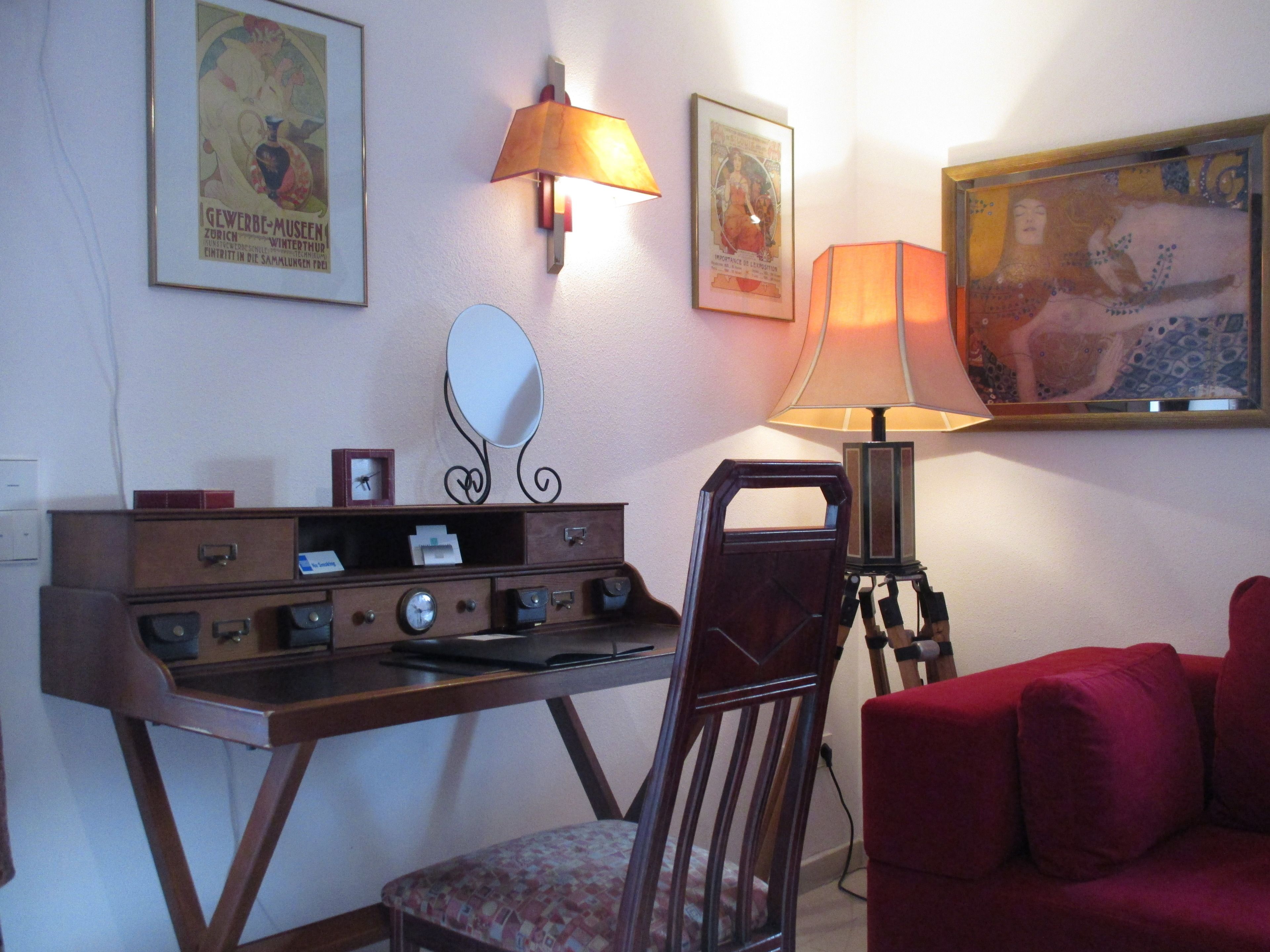 Une pièce avec un bureau en bois, une chaise, un canapé rouge, une lampe fixée au mur et des tableaux encadrés accrochés au mur.