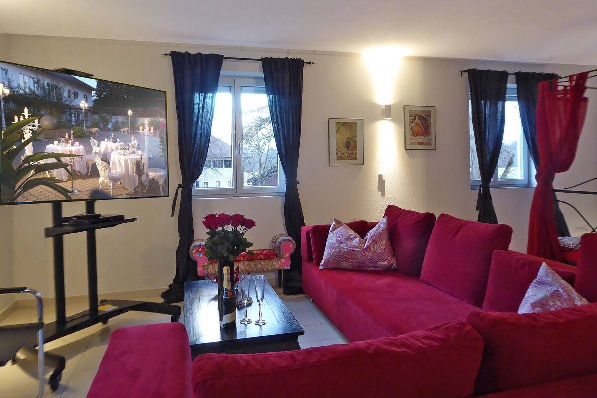 A living room with a red sofa, a coffee table with a vase of flowers, a TV on a stand, and a window with curtains.