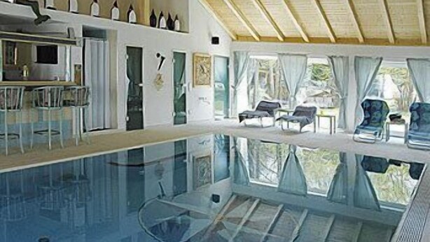 Una amplia piscina interior con paredes de vidrio transparente, techo de madera y una zona de estar con sillas y una mesa.
