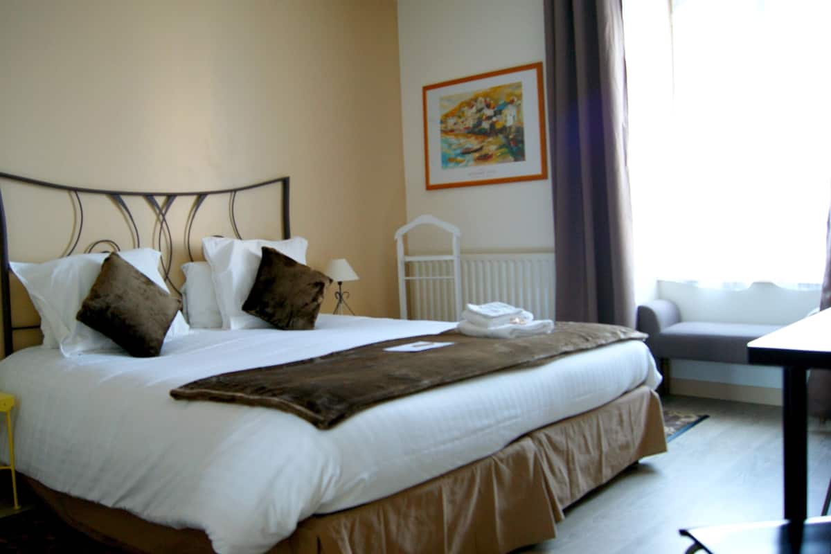 A neatly made bed with a brown headboard, white bedding, and a brown throw blanket. There is a white bedside table with a lamp and a phone. A white radiator and a painting are visible on the wall.