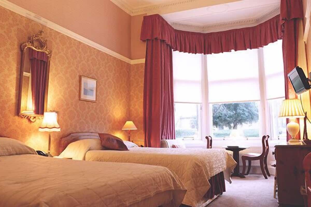 A hotel room with two beds, a large window with curtains, a desk with a lamp, and a chair.