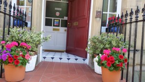 Una puerta roja con manija de latón, flanqueada por barandillas de hierro negro y macetas con flores rojas y púrpuras.