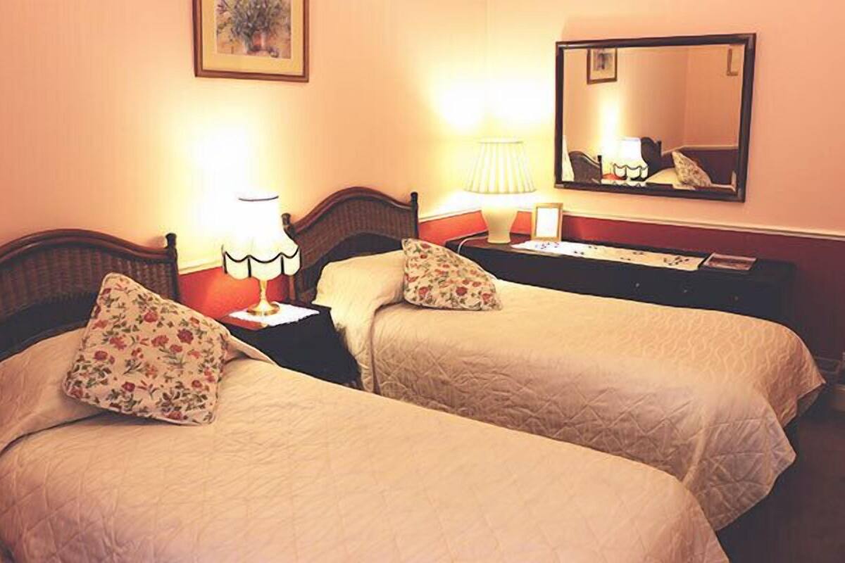 A hotel room with two beds, each with floral-patterned pillows, a bedside table with a lamp, and a mirror on the wall.