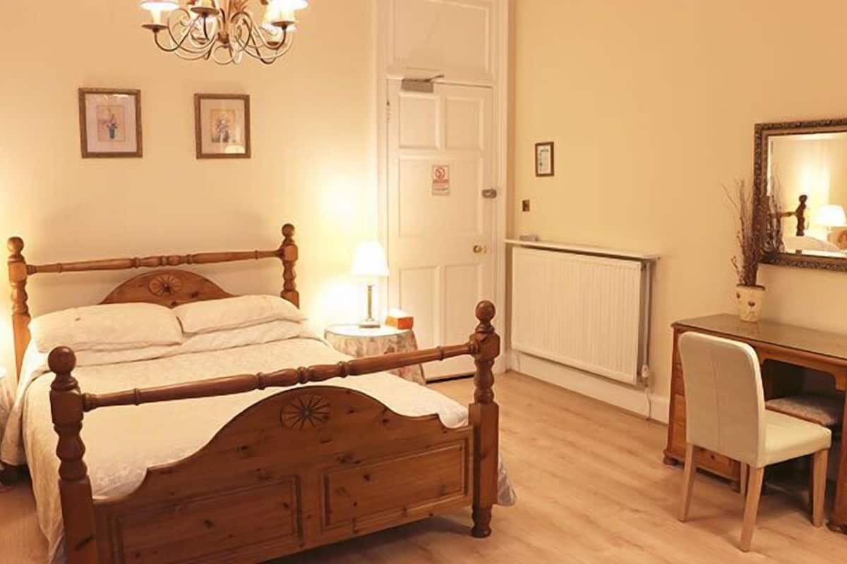 A bedroom with a wooden bed, a desk, and a chair. There is a radiator, a door, and two framed pictures on the wall.