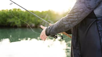 A person holding a fishing rod by a body of water with greenery in the background.